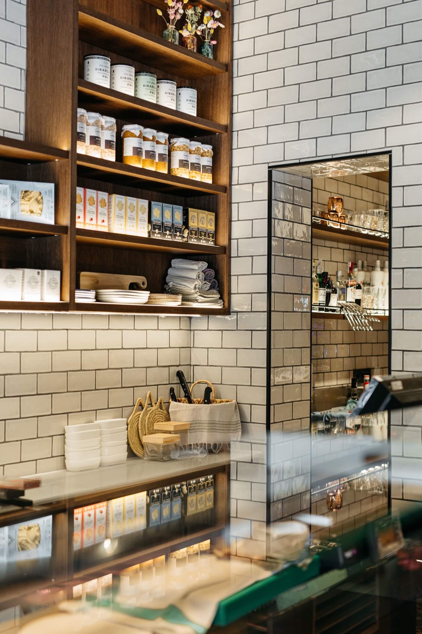 Estantería con productos gourmet y vajilla en el interior del restaurante Casa Petra en Barcelona, con revestimiento de azulejos blancos.