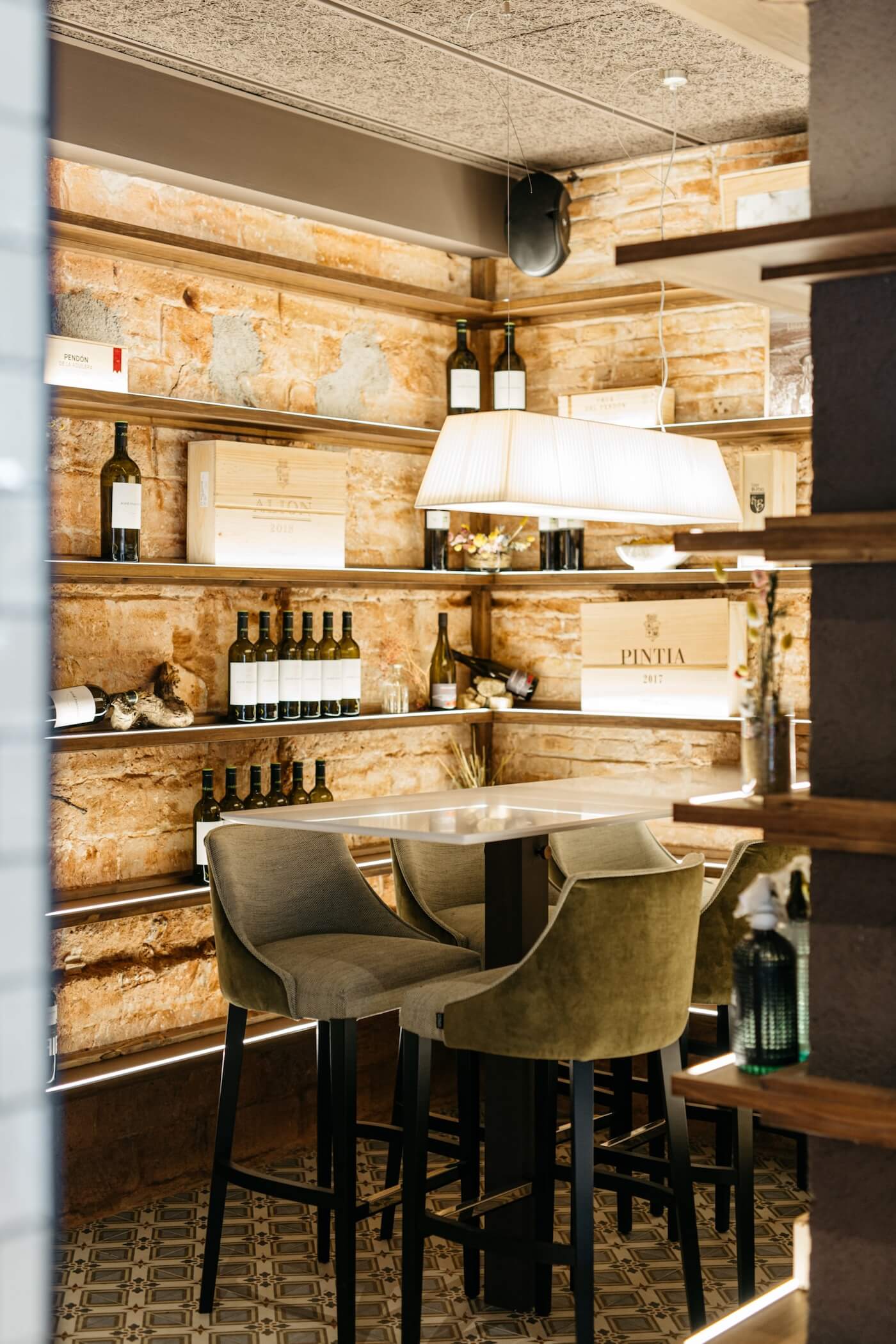 Zona de vinos en el interior del restaurante Casa Petra, con estanterías de madera, pared de ladrillo visto y lámpara colgante.