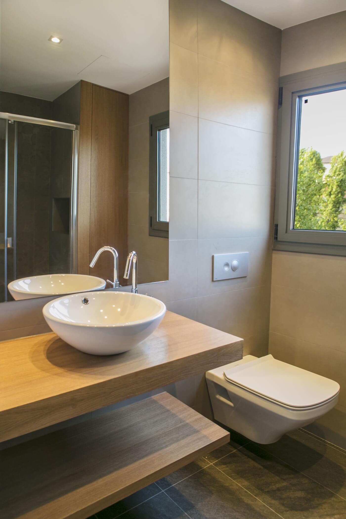Baño reformado con mueble de madera, lavabo sobreencimera, revestimiento cerámico y ventana con luz natural en vivienda de Sabadell.