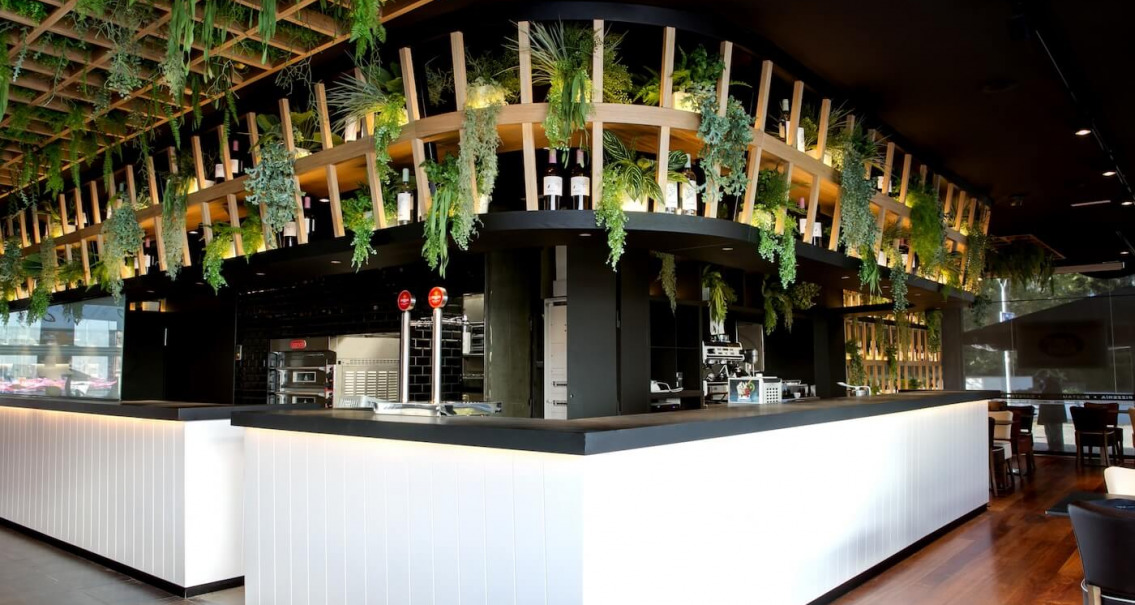 Barra del restaurante Crep Nova con estructura vegetal suspendida, botellas decorativas y diseño escenográfico.