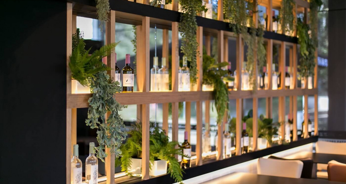 Estantería de madera con botellas iluminadas y vegetación colgante en restaurante Crep Nova, proyecto de interiorismo comercial.