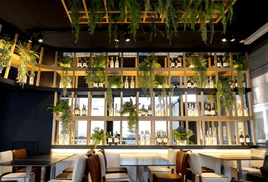 Diseño interior de restaurante con estantería de botellas retroiluminada, vegetación colgante y mobiliario a medida.