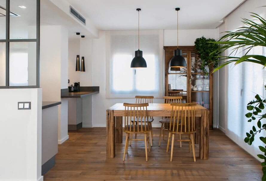 Comedor reformado con mesa de madera maciza, sillas estilo escandinavo, lámparas negras colgantes e iluminación natural.