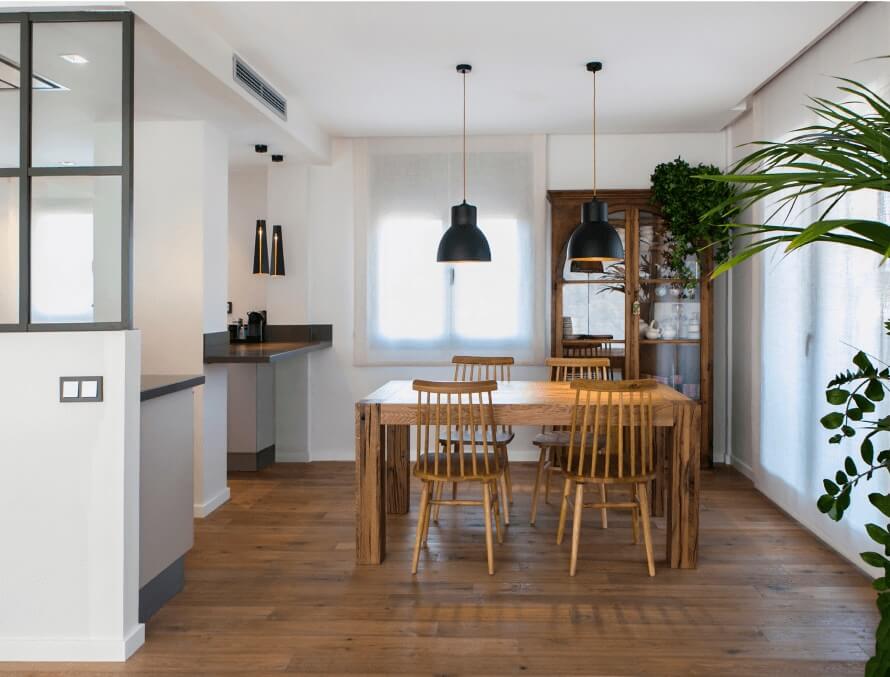 Comedor reformado con mesa de madera maciza, sillas estilo escandinavo, lámparas negras colgantes e iluminación natural.