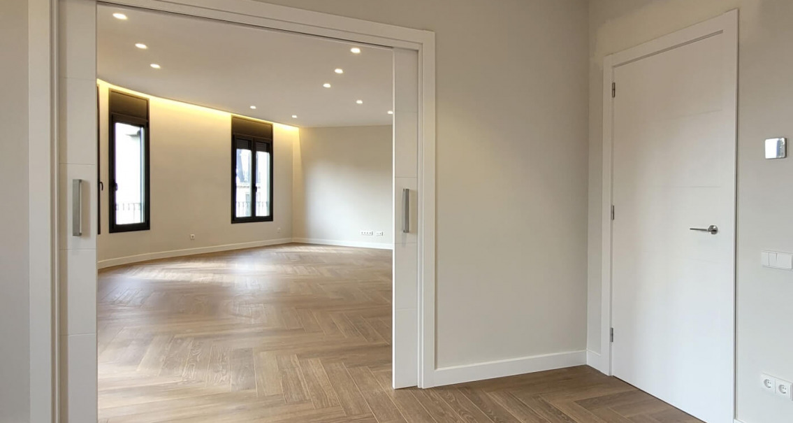 Vista desde una estancia hacia el salón con puerta corredera abierta, parquet en espiga e iluminación perimetral LED en reforma de piso.