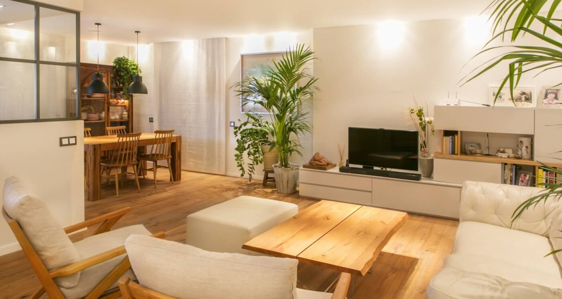 Salón comedor reformado con suelo de madera, mobiliario natural, iluminación cálida y decoración vegetal en vivienda de Sabadell.