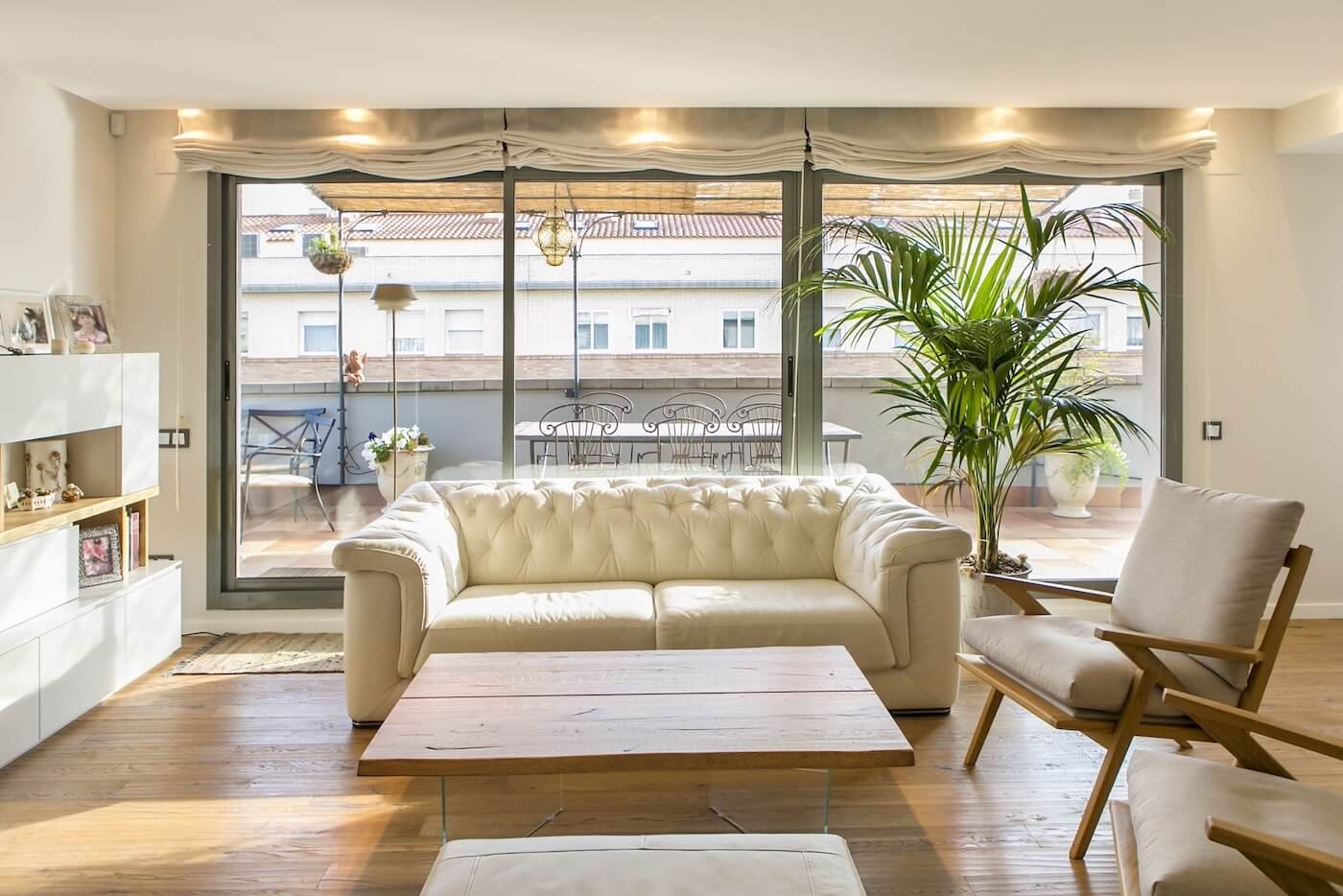 Salón reformado con sofá de piel blanca, suelos de madera natural, grandes ventanales y acceso a terraza con mobiliario exterior.