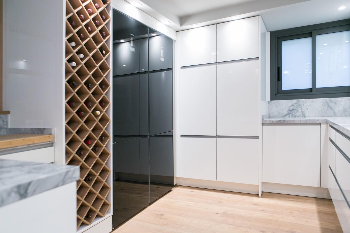 Detalle de vinoteca de madera integrada en una cocina moderna con mobiliario blanco y encimeras de mármol.